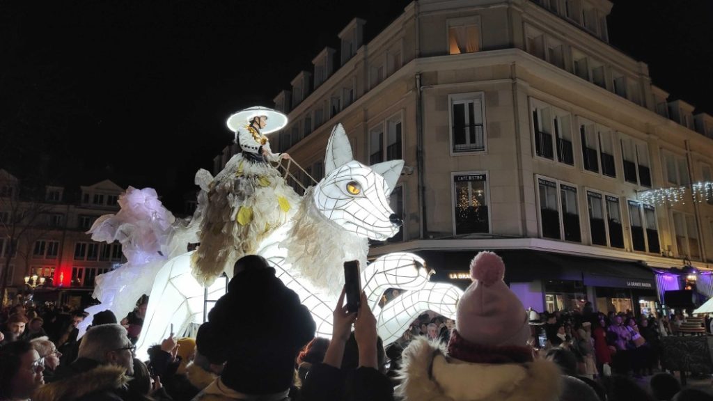 Photo of a lady with a large luminous hat mounted on a kind of immense fox, entirely illuminated from the inside by a white light.