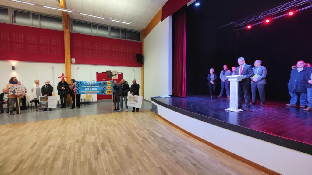 Photo of a wide shot of the Savignies village hall with, on the left, activists against the development of the airport and on the right the mayor, on the stage, speaking.